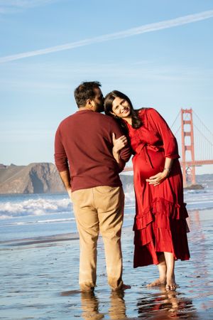 A couple shares a tender moment on Baker Beach, the pregnant woman in red cradling her bump near the Golden Gate Bridge.