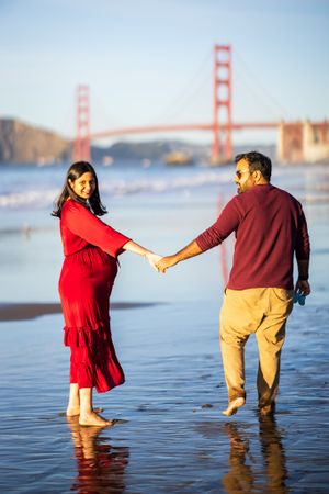 Pregnant couple holding hands on Baker Beach with Golden Gate Bridge backdrop, woman in red dress, sunny day.