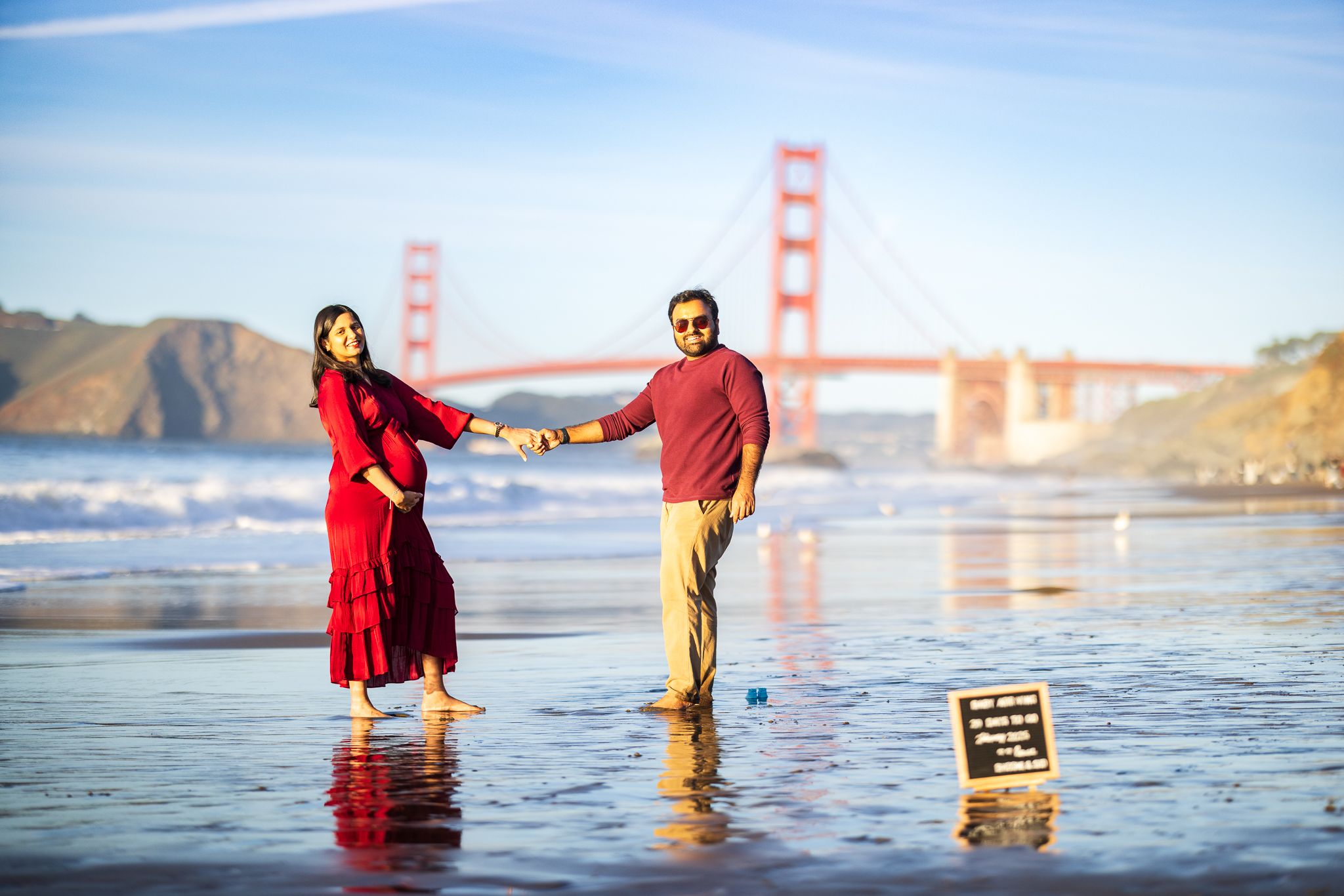 Expecting couple dancing on Baker Beach, Golden Gate Bridge visible, woman in red dress, maternity announcement sign.
