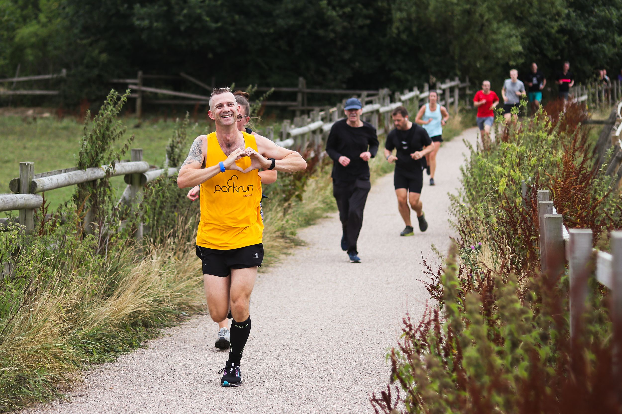 Man running at Woodgate Valley Country Park parkrun, making a heart with his hands and smiling, with runners in the background.
