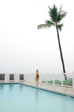 Woman in black bikini standing by a rooftop pool with a tall palm tree and misty ocean view.