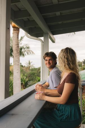 A smiling couple leans on a porch railing with tropical palm trees and lush greenery visible in the background.