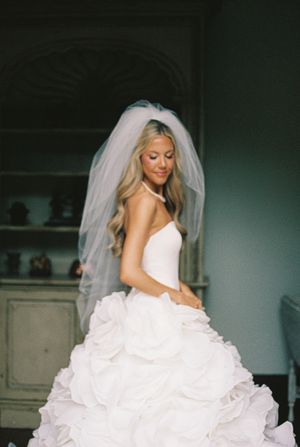 Elegant bride in a white ruffled ball gown with veil, long blonde hair, posing gracefully on her wedding day.