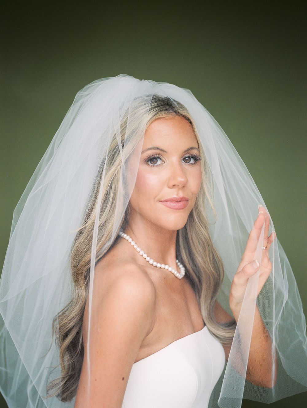 Bride wearing a white veil and pearl necklace with elegant makeup and flowing blonde hair against a green background.