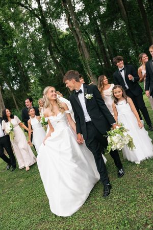 Bride and groom walk hand-in-hand with wedding party in white and black formal attire on lush green lawn.