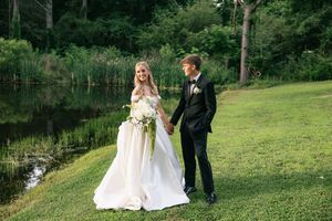 Bride and groom holding hands on green lawn near pond, bride in white gown with bouquet, groom in black suit.