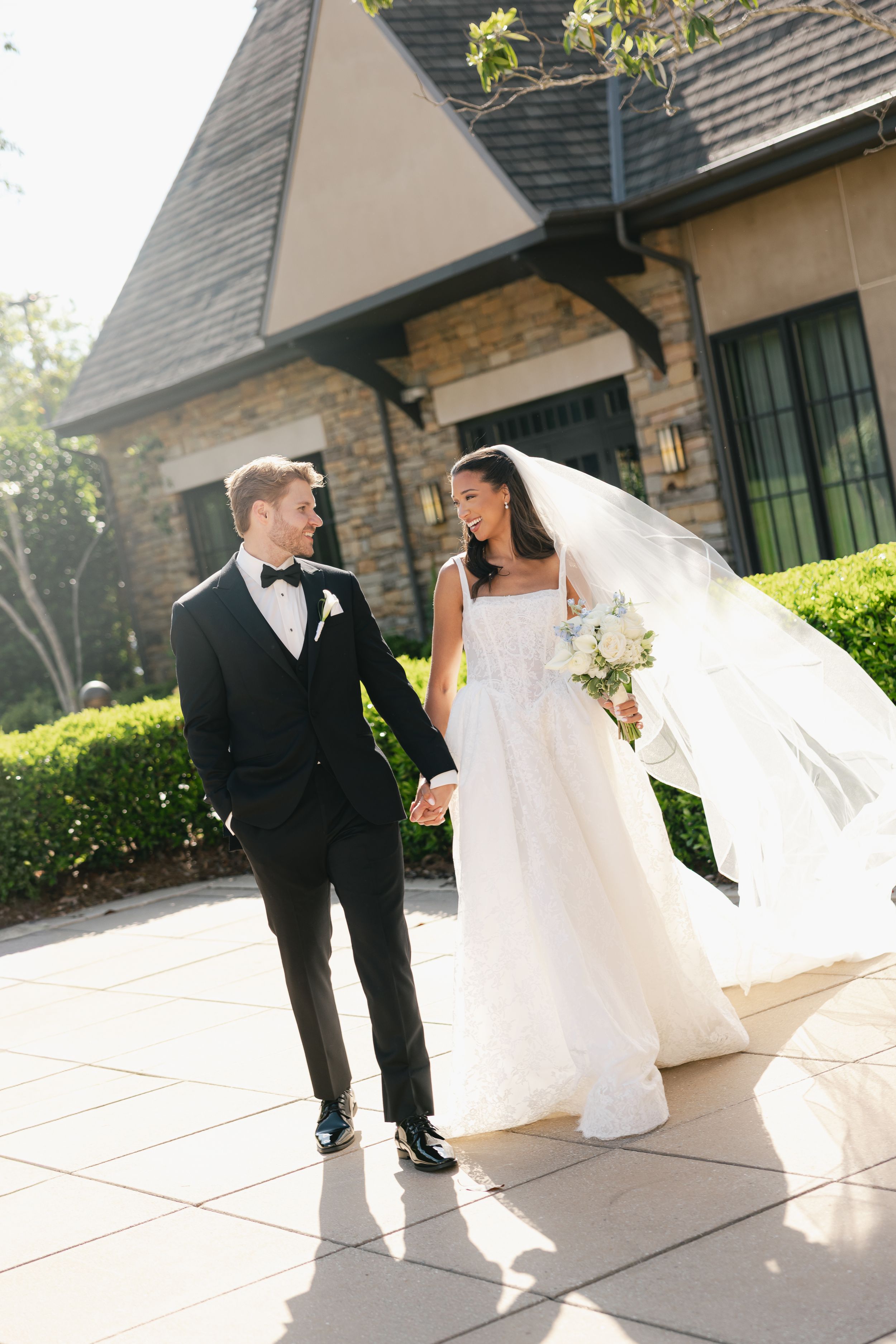 Bride and groom holding hands walking outside stone building, bride in white gown with veil, groom in black tuxedo.
