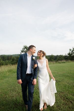 A laughing bride and groom walk hand in hand through an open green meadow under a cloudy sky.