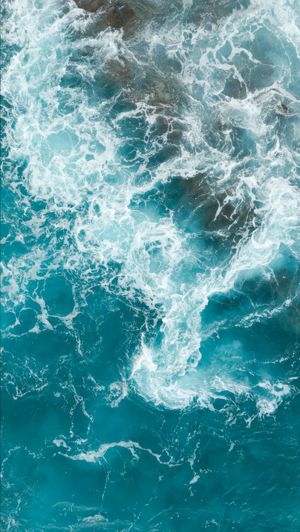 Aerial view of turbulent turquoise ocean waves with white sea foam swirling on the water surface.
