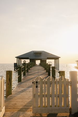 Wooden pier with white gate leading to covered boathouse over calm ocean waters at golden sunset.