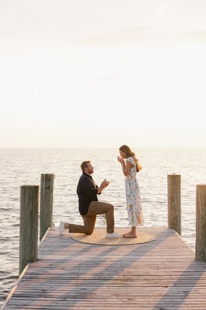 Man proposes on bended knee to woman on a wooden dock at sunset over calm ocean waters.