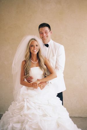 Smiling bride and groom posing together on their wedding day, bride in white gown with veil, groom in white tuxedo jacket.