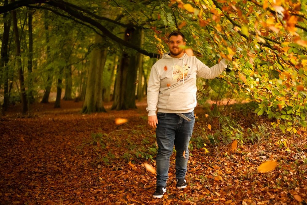 A person in a light colored hoodie walks through a forest path covered in autumn leaves.