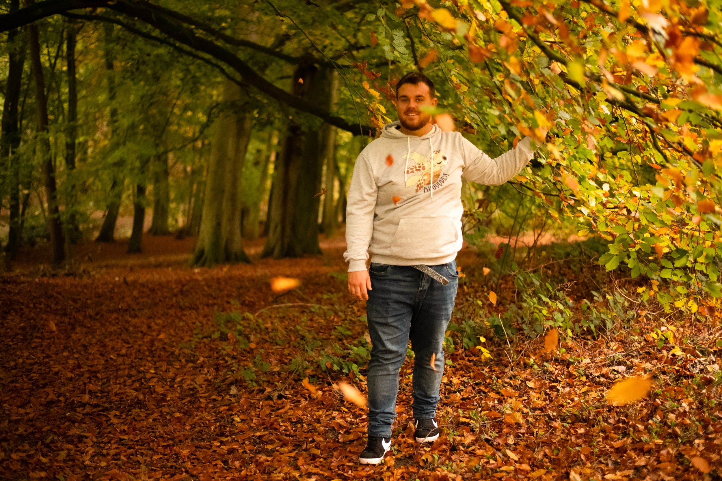 A person in a light colored hoodie walks through a forest path covered in autumn leaves.