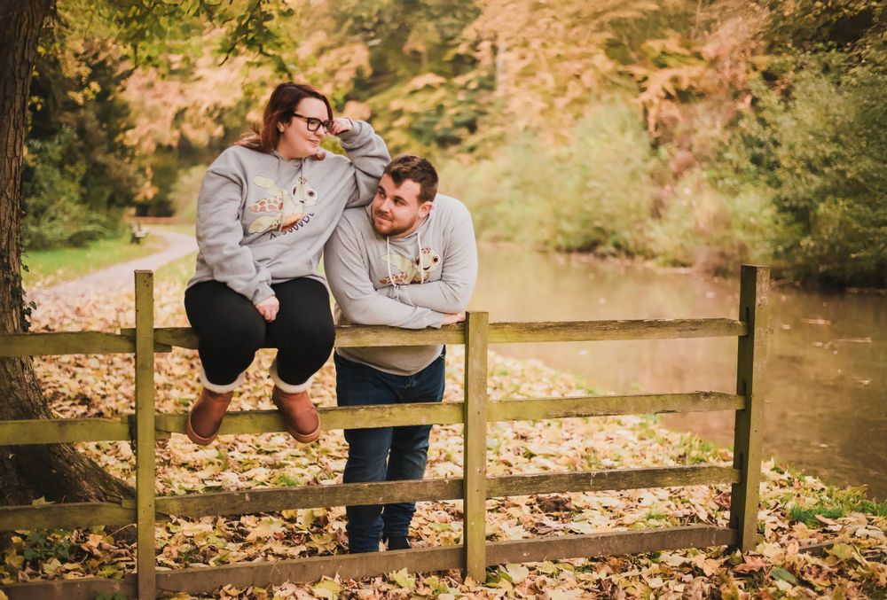 A couple in coordinated outfits sits on a wooden fence surrounded by fall foliage.