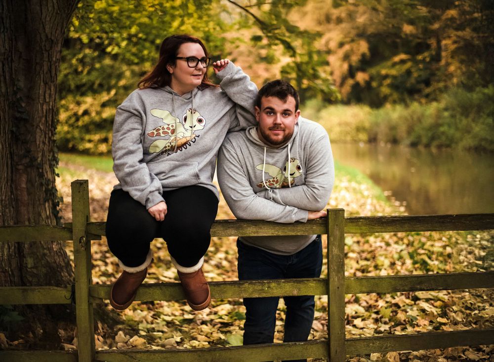 A couple wearing matching gray sweatshirts sits on a wooden fence by a pond in fall.