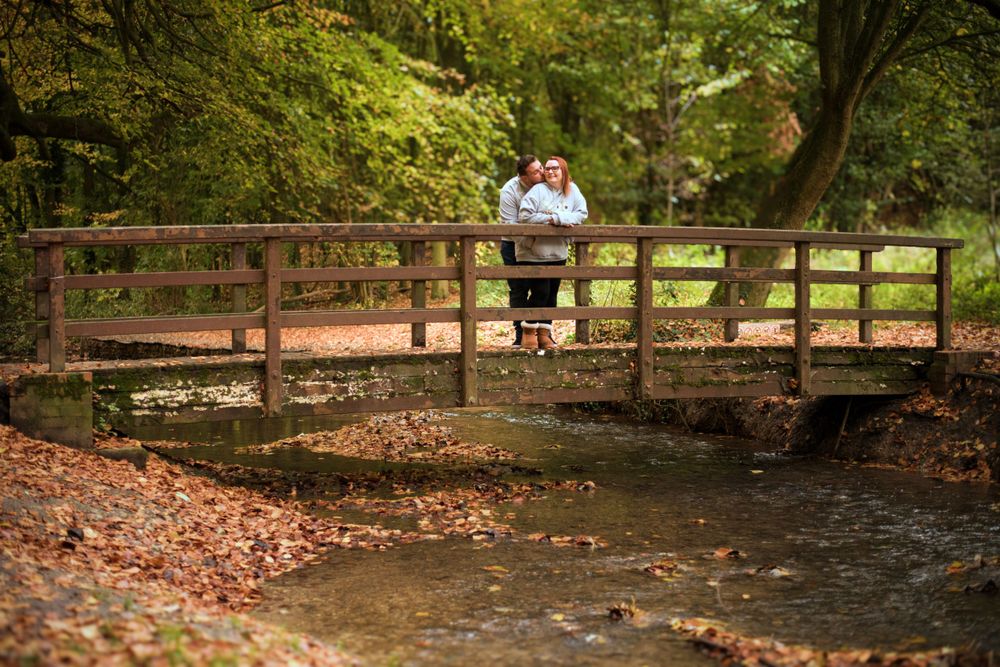 Two people stand on a wooden bridge over a creek surrounded by fallen autumn leaves.