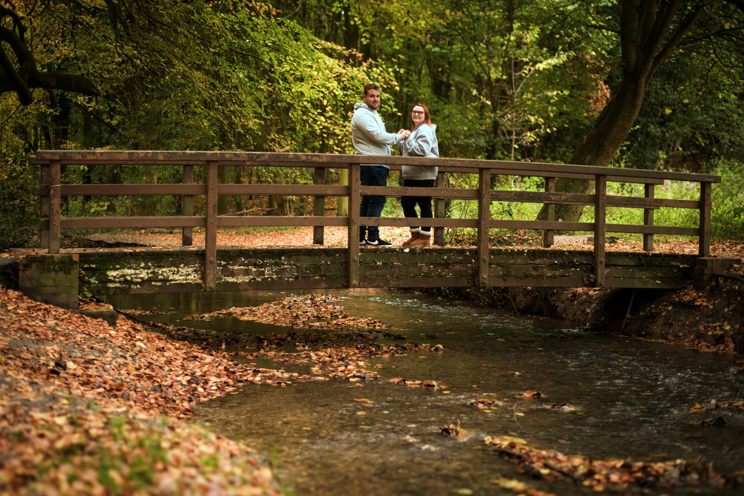 A couple stands on a rustic wooden bridge over a creek in an autumn forest setting.