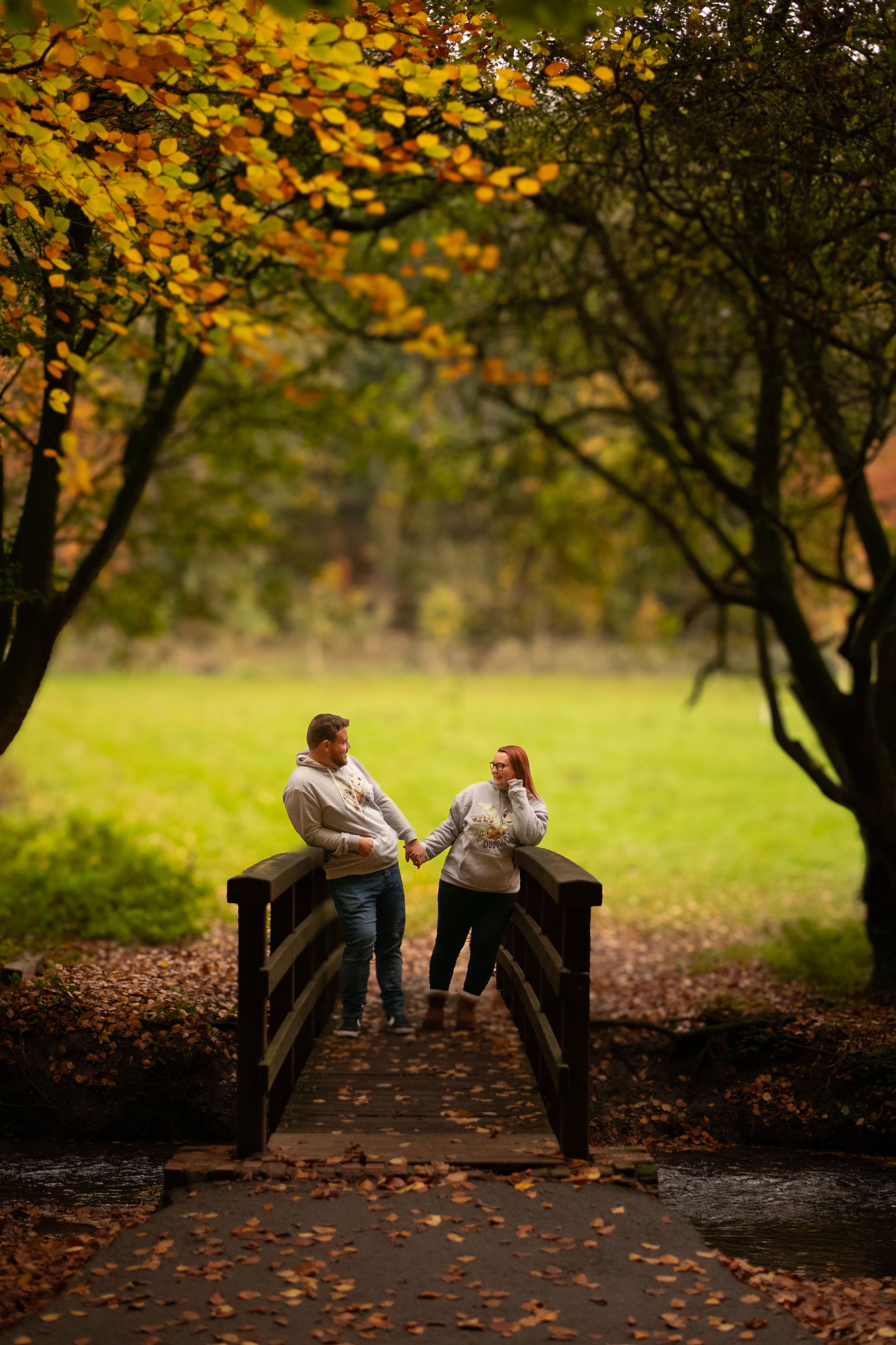 Two people share a moment on a wooden bridge framed by golden autumn trees.