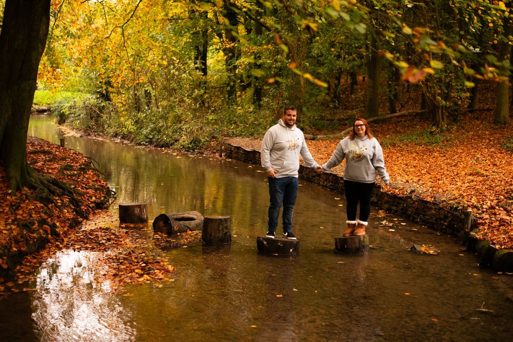 Two people stand on stones in a creek while holding hands in an autumn forest.