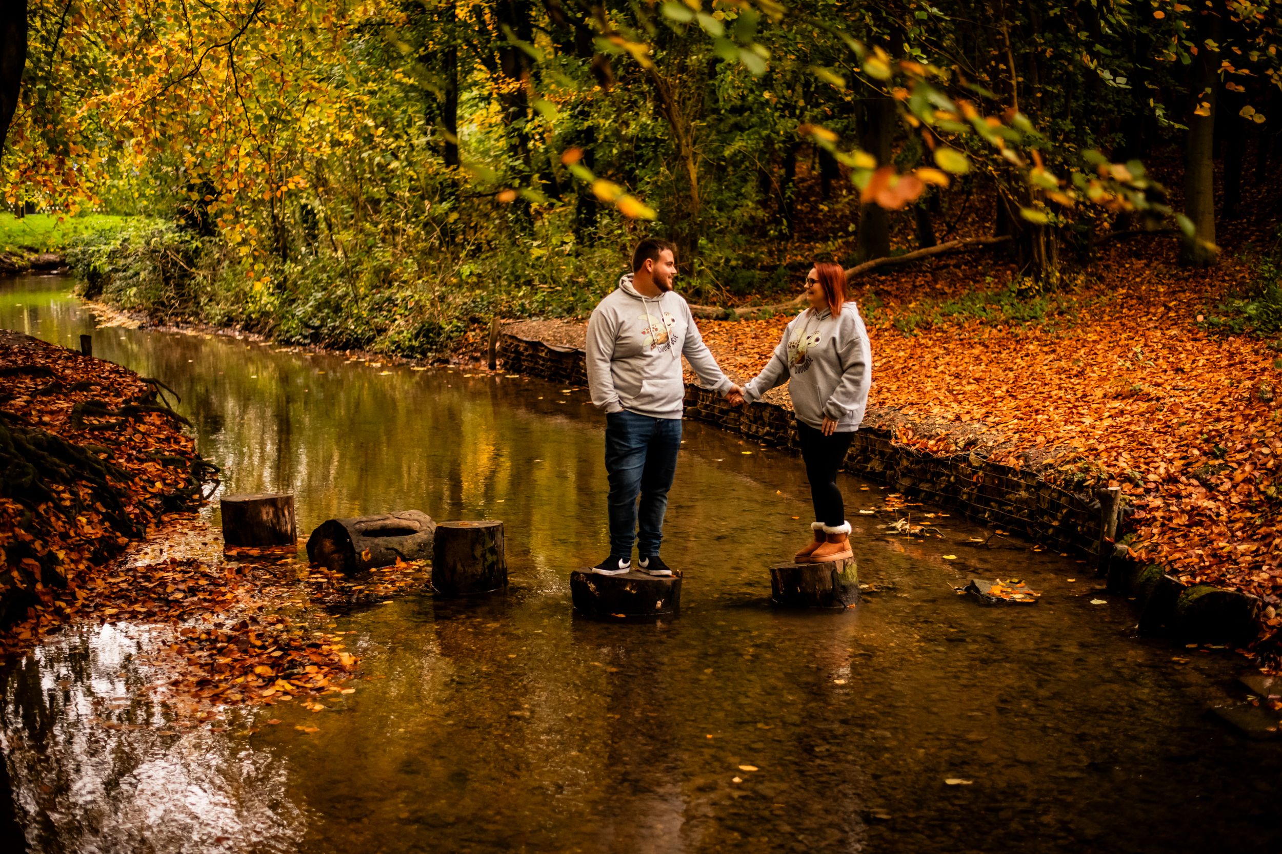 A couple balances on stepping stones across a creek surrounded by fall foliage.