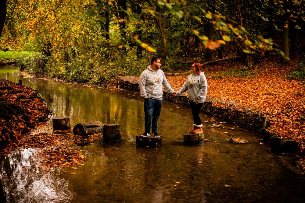 A couple balances on stepping stones across a creek surrounded by fall foliage.