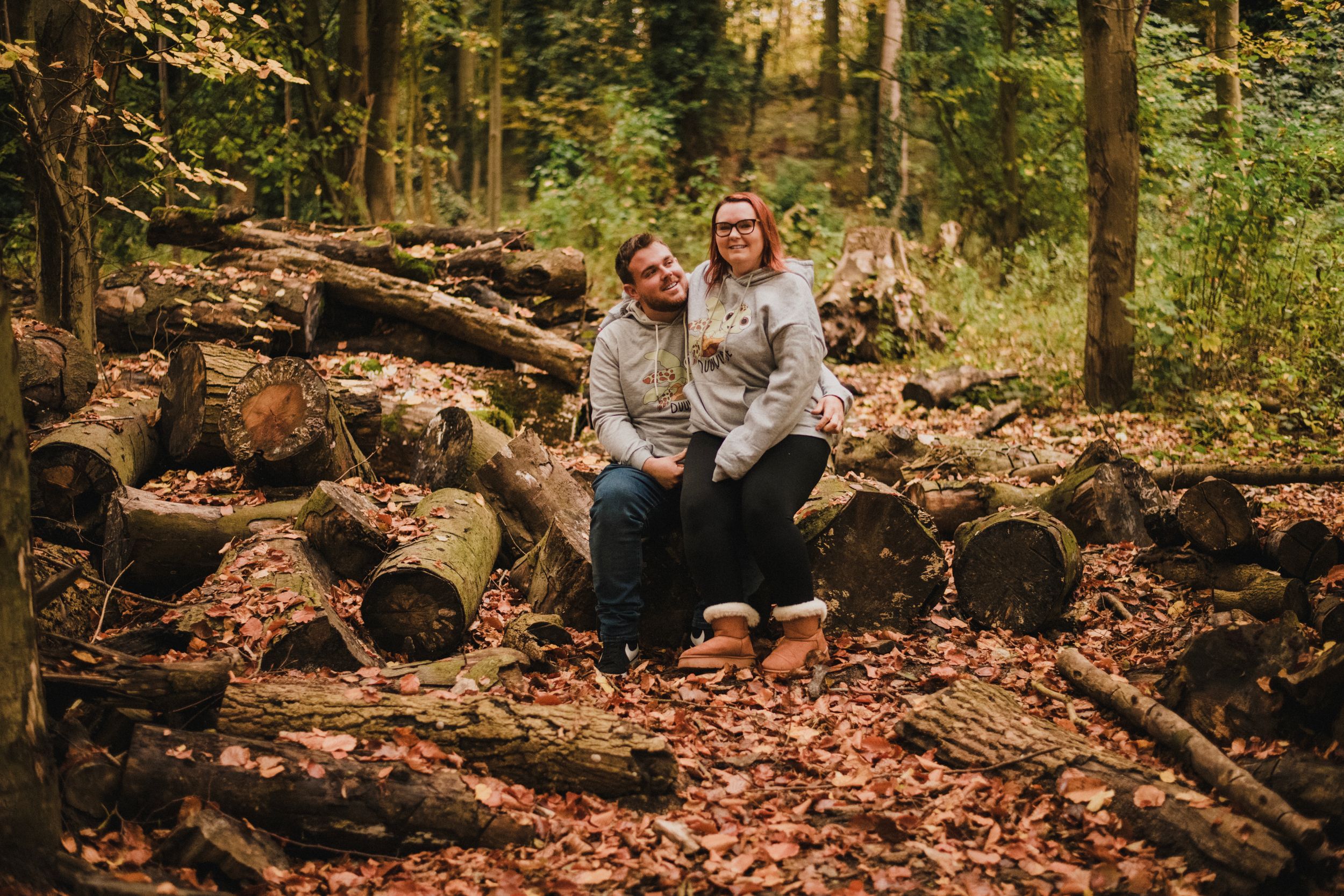 Couple sitting together on rustic log pile surrounded by autumn leaves in a forest setting.