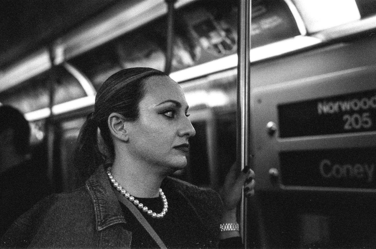 A black and white photo of a passenger on a subway train wearing a pearl necklace and denim jacket.