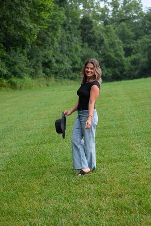 Woman in casual attire walking and posing on a grassy hillside surrounded by trees.