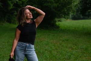 Woman in black top and jeans poses playfully in a green grassy field on a sunny day.