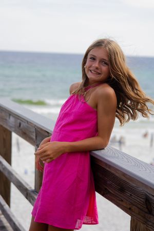 A young girl in a bright pink dress smiles while posing on a beachside boardwalk.