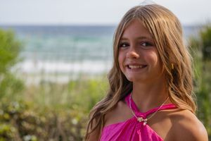 A girl in a pink halter dress smiles with the ocean and beach visible in the background.