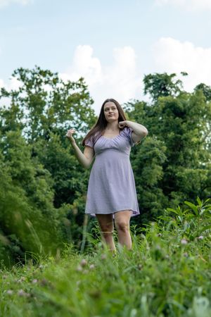 A figure in a light purple dress stands in a lush green field on a sunny day.