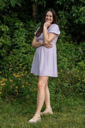 Someone wearing a light-colored dress poses casually in front of flowering bushes.
