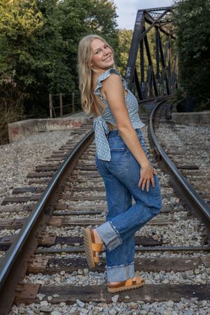 Person in denim overalls and blue top poses playfully on railroad tracks.