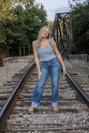 Person wearing blue polka dot top and jeans poses playfully on rustic railroad tracks with metal bridge in background.
