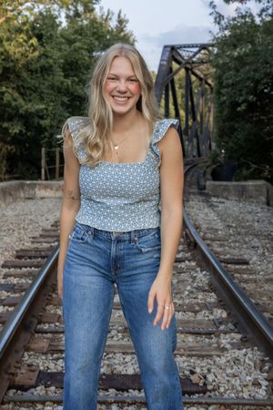 Person in casual outfit stands on railroad tracks surrounded by greenery and a metal bridge structure.