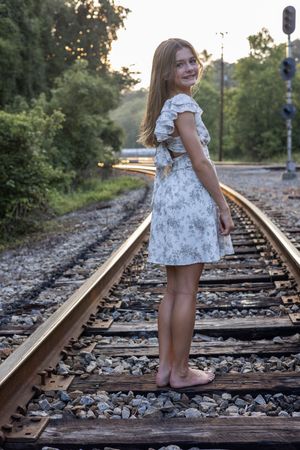 Person in light summer dress poses along railroad tracks at sunset with trees in background.