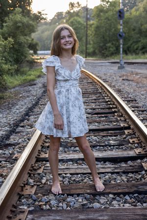 Figure in white patterned dress strikes playful poses on railroad tracks during evening photoshoot.
