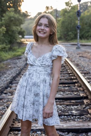 Person wearing white floral dress poses on railroad tracks with natural lighting and greenery.