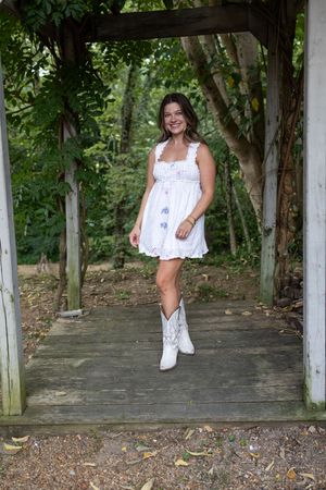 Person in white eyelet top poses under wooden pergola surrounded by lush green trees.