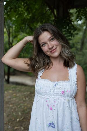 A woman in a white summer dress poses playfully on a wooden deck surrounded by greenery.
