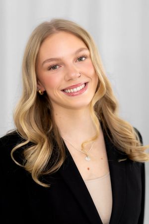 Professional headshot portrait of a person with blonde wavy hair wearing a black blazer and delicate necklace against light background.