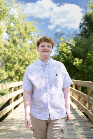 Young person in white polka dot shirt poses on wooden bridge in sunlit setting.