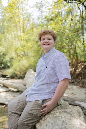 A young person in a polka dot shirt and khaki pants sits casually on a large rock outdoors on an autumn day.