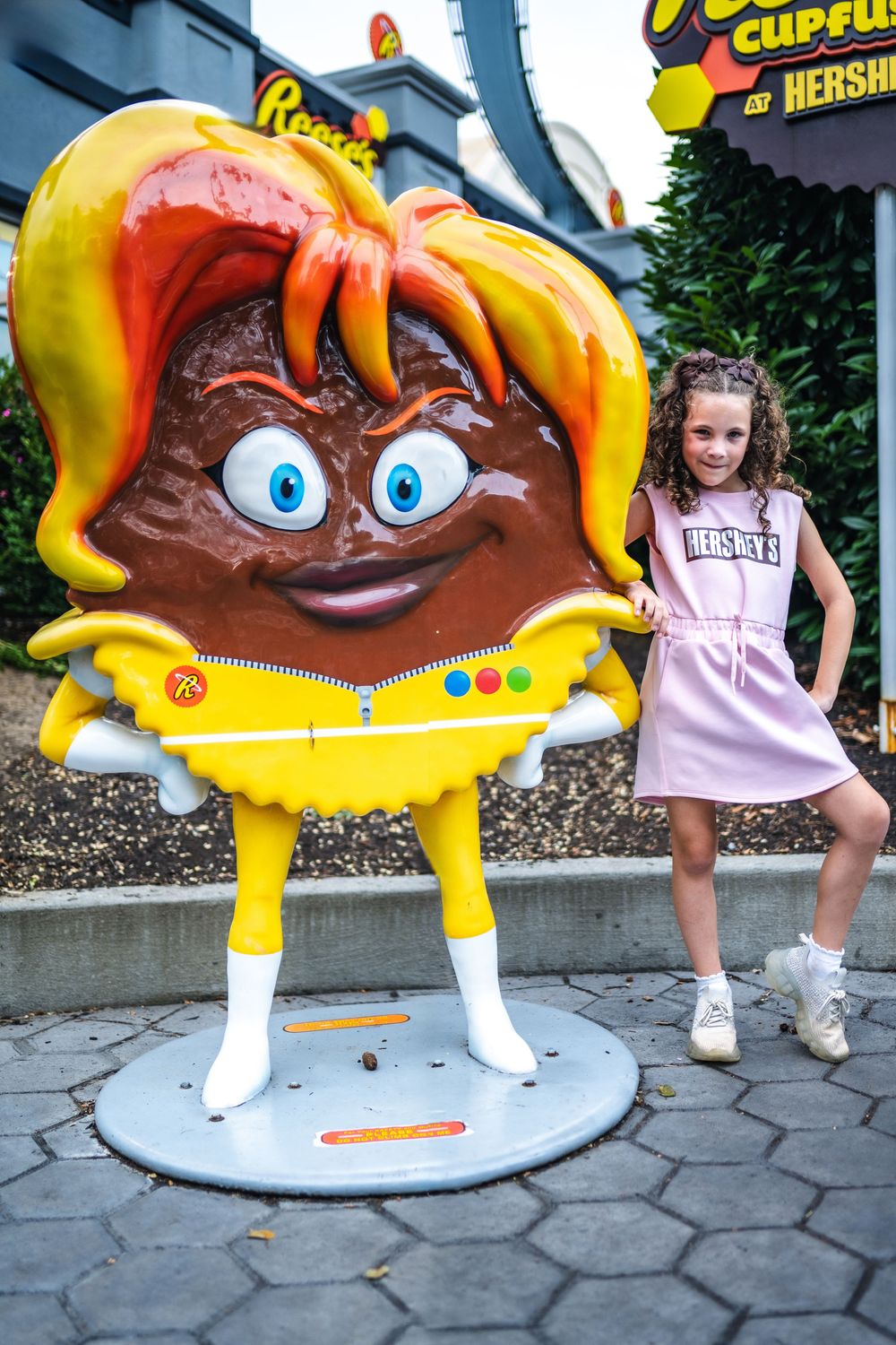 A large Reese's Peanut Butter Cup mascot statue in a yellow dress stands at Hersheypark near a young visitor.