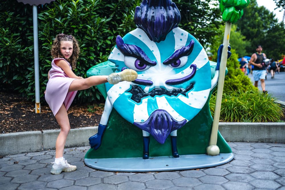 A young child in casual attire poses playfully with a large blue and white decorative flower sculpture in an outdoor setting.