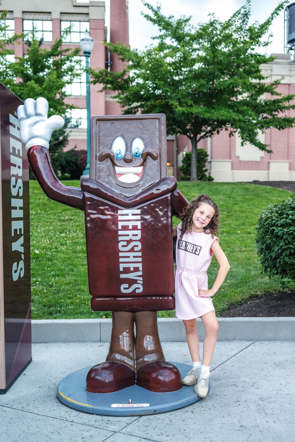 A large Hershey's chocolate bar mascot statue stands next to someone posing by landscaping outside a building.