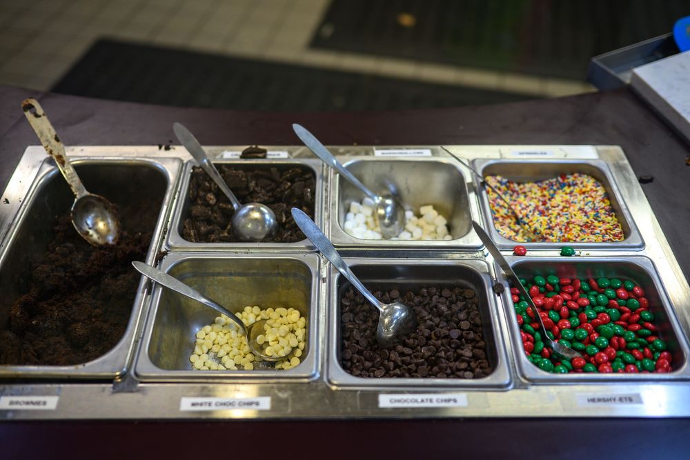 Metal containers filled with various ice cream toppings including chocolate chips, sprinkles, and candies at a dessert station.