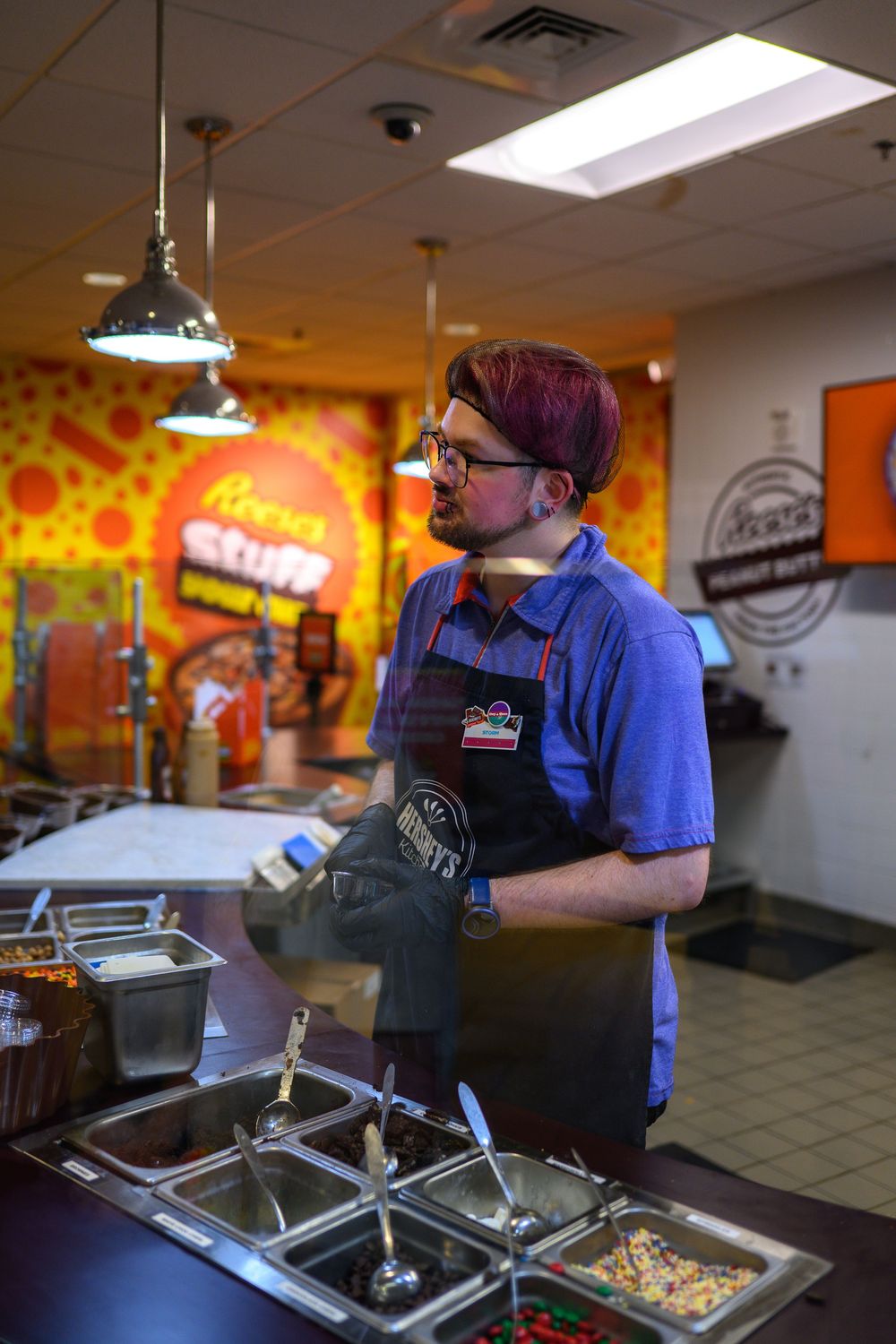 Fast food restaurant worker preparing ingredients at stainless steel prep station under fluorescent lighting.